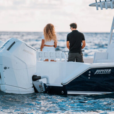 Two people stand at the stern of a white Jupiter boat, facing the open ocean. The outboard engine is visible on the left, and the boat has a sleek design.