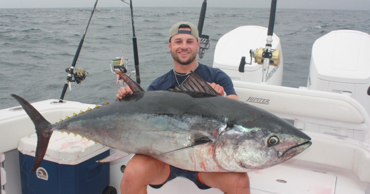 A man is sitting on a boat, smiling proudly while holding a large, freshly caught tuna. The boat is equipped with fishing rods and other gear, and the ocean is visible in the background, appearing slightly choppy under a cloudy sky. The man is wearing a cap backwards and casual clothing, emphasizing the triumphant moment of his successful fishing trip.