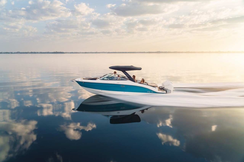 Image of a sleek blue and white motorboat speeding across a calm, reflective body of water during a serene sunset. The boat's wake creates gentle ripples, and the sky is filled with soft, scattered clouds, mirrored beautifully on the water's surface. Two people are seen relaxing on the boat, enjoying the peaceful atmosphere.