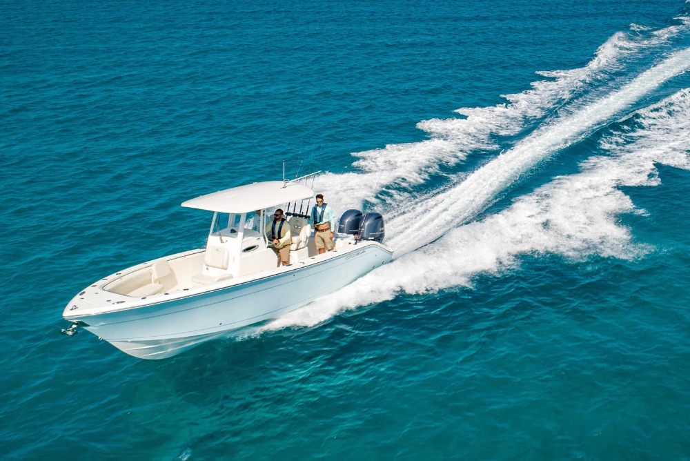 Cobia 262CC speeding across clear, blue ocean waters. The boat has a T-top roof and is equipped with two powerful outboard engines. Two individuals are standing under the roof, facing forward, with one person appearing to be steering the boat. The boat leaves a white, foamy wake behind it as it cuts through the water, indicating high speed. The scene conveys a sense of adventure and enjoyment on the open sea.