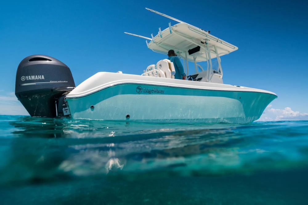 EdgeWater 245CC floating on clear, calm water under a bright blue sky. The boat is equipped with a Yamaha four-stroke outboard motor. A person wearing a blue shirt and hat is standing at the helm under a white canopy. The water reflects the boat's hull, enhancing the tranquil and vibrant maritime scene.