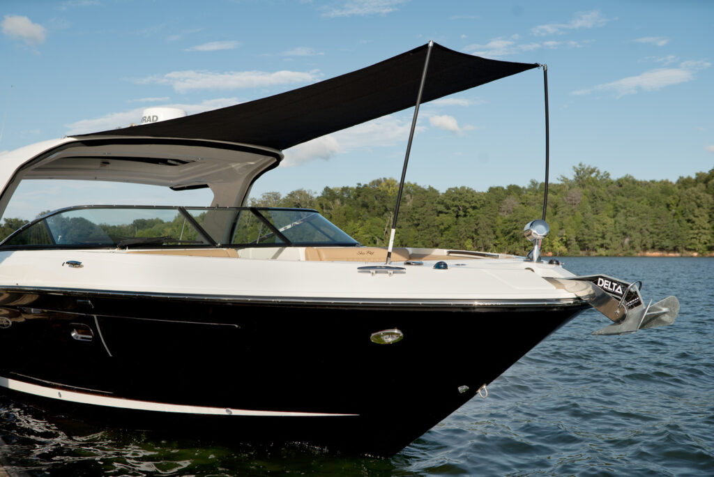 A luxury motor yacht with a black hull and white superstructure is anchored on a calm lake. The boat features a sleek, modern design with a large bow area covered by an extended black sunshade, providing ample shade and comfort for passengers. The name "Sea Ray" is visible on the side of the boat. The background includes lush green trees and a clear blue sky with a few clouds.