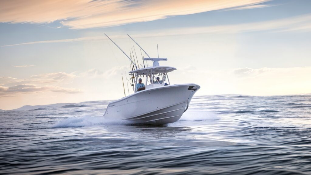 Image of a Cobia fishing boat with multiple fishing rods mounted on the back, speeding across the water at sunset. Two people are visible on board, with the boat cutting through gentle waves under a pastel-colored sky.