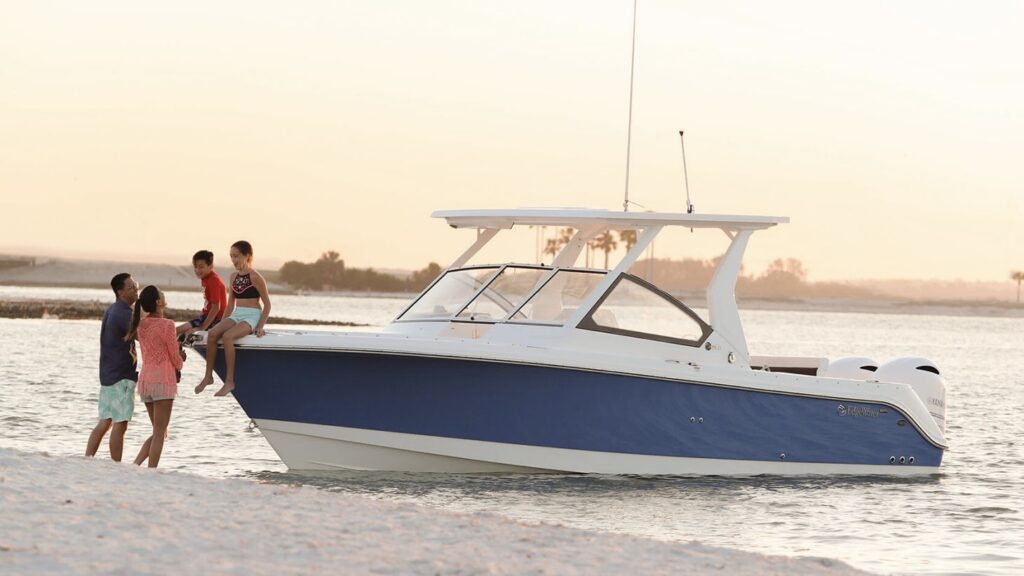 Image of an EdgeWater boat anchored near a sandy beach at sunset. A family of four, including two children sitting on the bow, is seen enjoying their time together. The sky is softly lit with warm hues, highlighting a serene and picturesque moment.