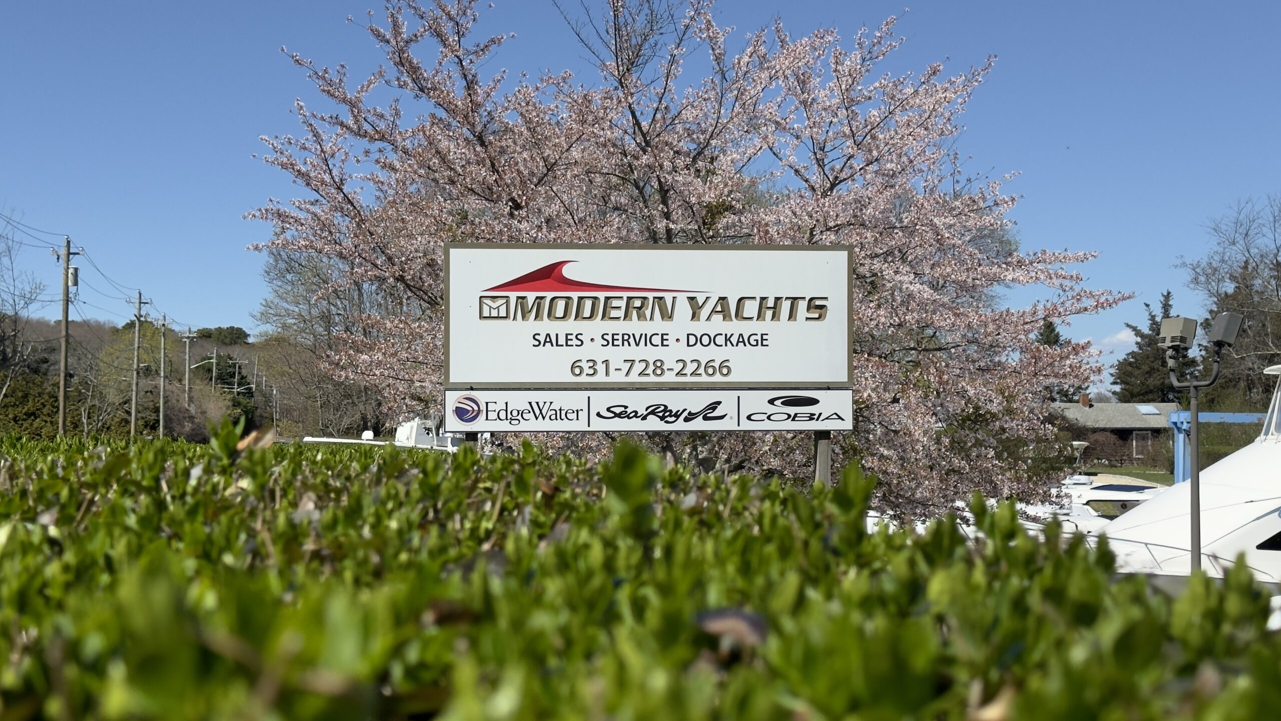 Image of a Modern Yachts sign with the text 'Sales • Service • Dockage' and a phone number (631-728-2266) prominently displayed. The sign also features logos for EdgeWater, Sea Ray, and Cobia, positioned at the bottom. The background includes blooming cherry blossom trees and a clear blue sky.